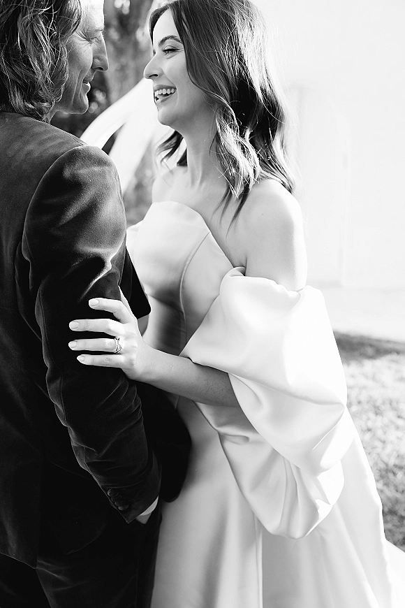 Couple portrait in a black and white wedding photo, bride hugging groom in a dark suit, veil and ring visible on an outdoor lawn with trees