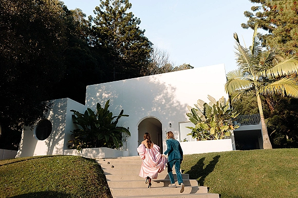 Couple portrait of bride and groom walking up stairs, groom holding her pink gown by a modern white villa with palm trees and blue sky