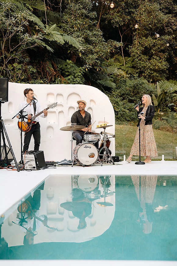 Wedding live band setup with microphones, electric guitar and drum kit on a poolside stage with string lights and white backdrop wall