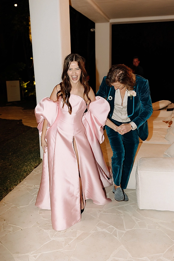 Reception exit moment as bride in a pink strapless puff-sleeve gown with high slit walks with groom in teal velvet suit on a patio at night