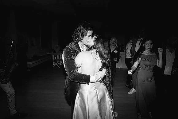 Wedding kiss moment during first dance kiss on a dim reception dance floor, bride in backless gown and groom in velvet jacket as a saxophone player performs nearby