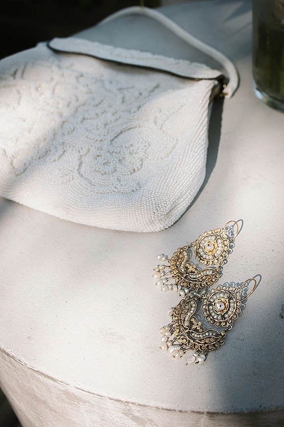 Bridal earrings with pearl drop earrings in gold filigree beside an embroidered beaded clutch on a stone tabletop near a glass vase