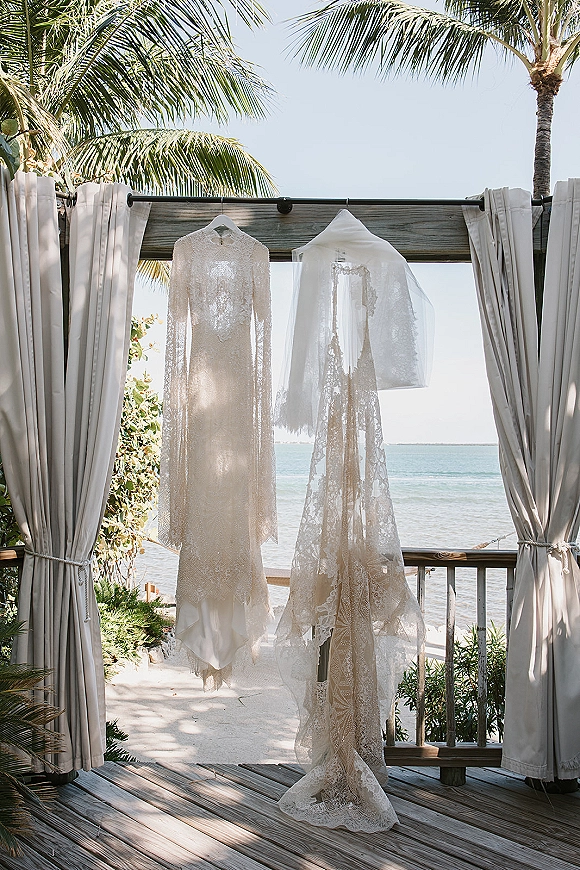 Wedding dress lace wedding dress hanging from a hanger with a bridal veil beneath canopy curtains on a deck overlooking palm trees and ocean
