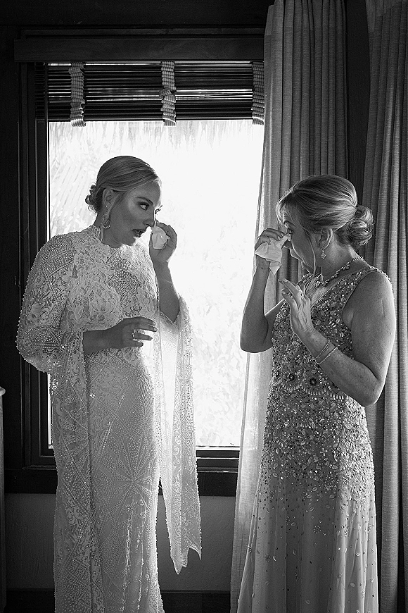 Getting ready moment as bride wipes tears while her mother comforts her by a window, lace gown and beaded cape, black and white candid