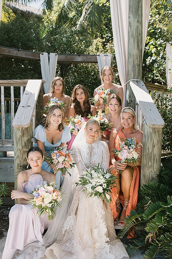 Bridesmaids portrait with bride in lace gown and veil on a wooden staircase, holding bouquets against sunlit tropical greenery