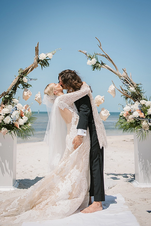 Wedding kiss at a beach wedding kiss under a driftwood floral arch with seashells, ocean and blue sky behind the couple