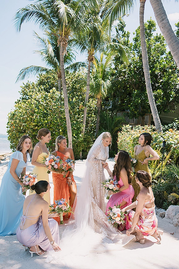 Bridesmaids group photo with bride with bridesmaids holding bouquets in colorful dresses on a sandy path by palm trees and ocean sunlight