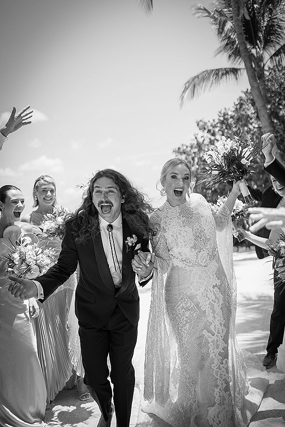 Recessional moment as bride and groom walk a sandy path, veil and bouquet in hand, wedding party cheering under palm trees in sunlight