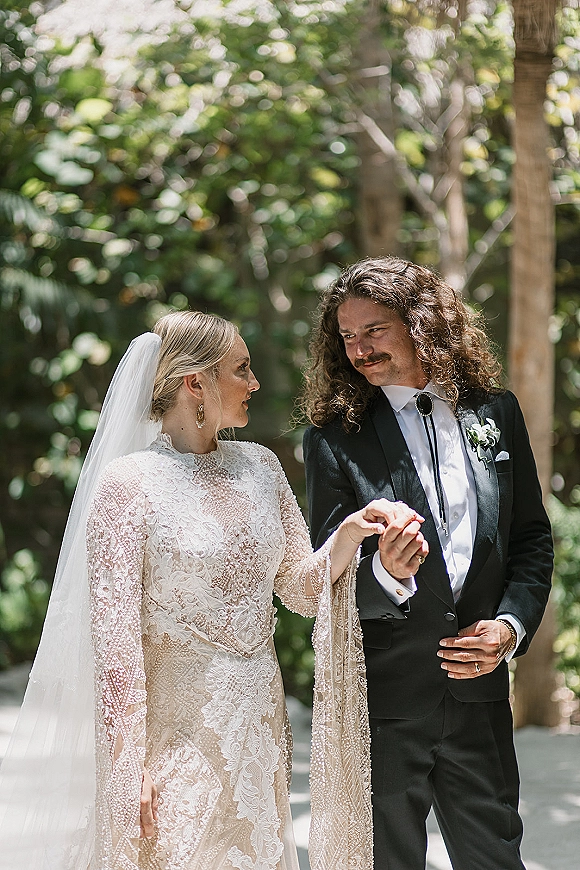 Couple portrait of bride and groom holding hands, her lace long-sleeve gown and veil, his tux with bolo tie in sunlit garden trees