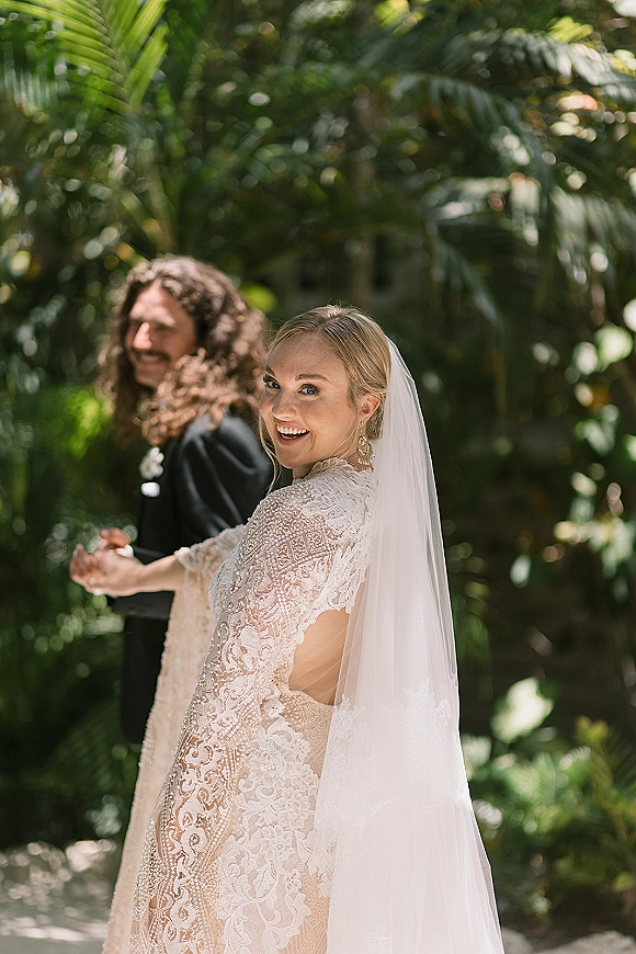 Wedding couple portrait of bride looking back while holding hands with groom, long veil trailing through tropical garden greenery with palm leaves