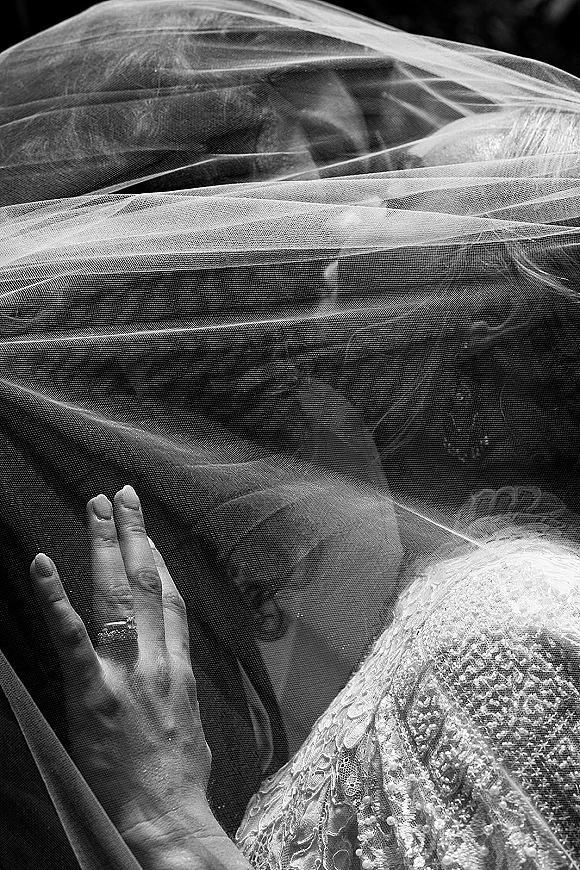 Wedding kiss portrait of bride and groom kissing beneath a sheer veil, her ringed hand on his face against a dark backdrop