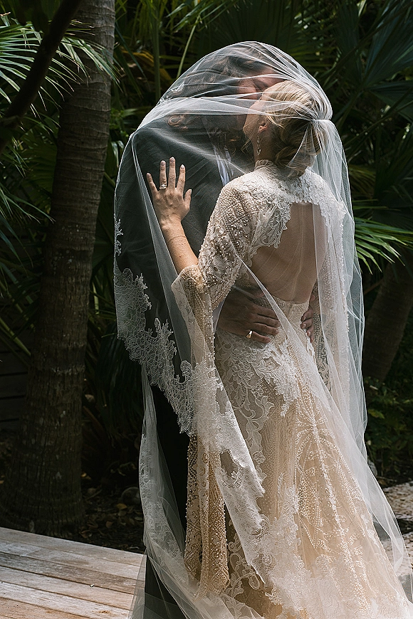 Wedding kiss portrait of bride and groom embracing under a veil, her lace long-sleeve open-back dress on a wooden deck with palm trees behind