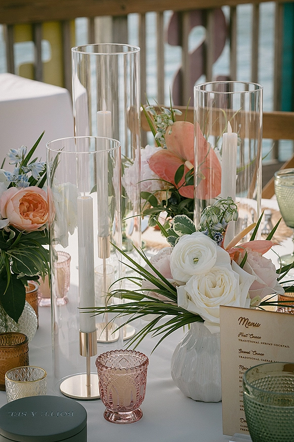 Reception tablescape with wedding table centerpiece of blush and white roses, anthuriums, greenery and hurricane candles on a waterfront deck