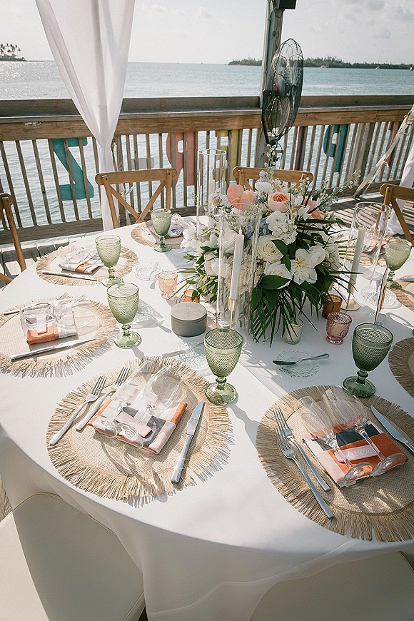 Reception tablescape with white tablecloth, rattan fringe placemats, green goblets, and pink votives on a waterfront deck by the ocean
