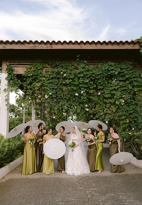 Bride with bridesmaids holding parasols and bouquets, bride in lace dress and veil under a vine-covered archway in a tropical courtyard walkway