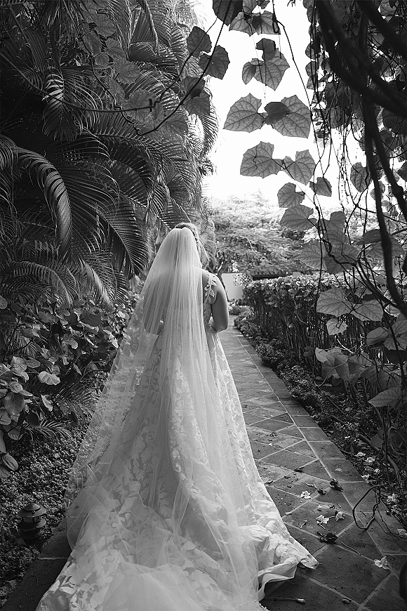 Bridal portrait of a bride from behind with a long wedding veil train over a lace dress, walking along a tropical garden stone path