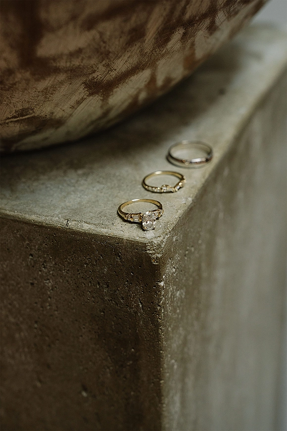 Wedding rings with an oval diamond engagement ring stacked on gold bands, arranged on a stone ledge beside a dried palm leaf