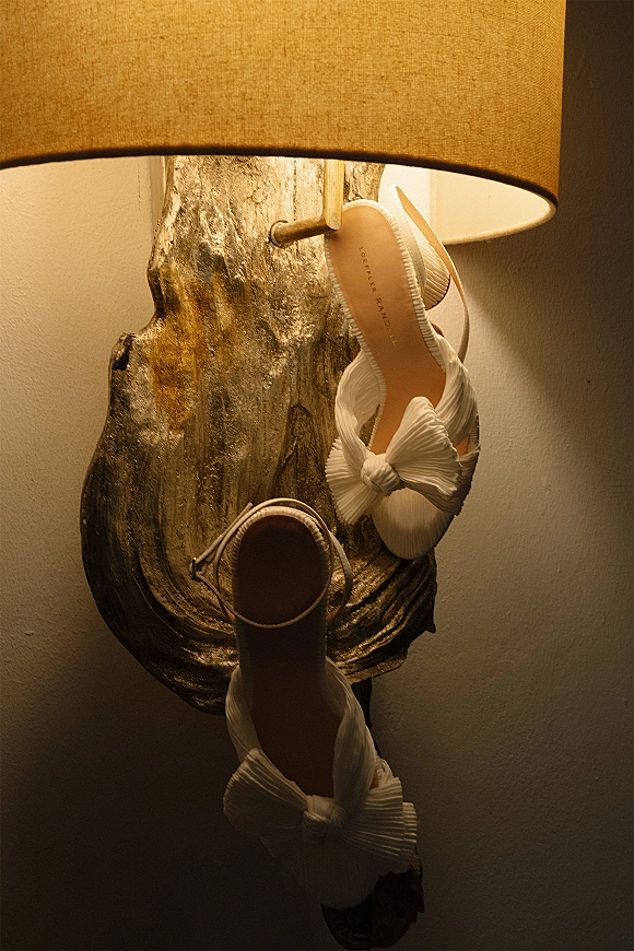 Bridal shoes in a wedding shoes flat lay, white heeled slingback sandals with bow detail beside a table lamp against a neutral wall