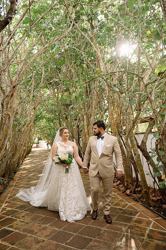 Couple portrait of bride and groom walking hand in hand, bride holding bouquet on a sunlit tree-lined brick path beside a white wall