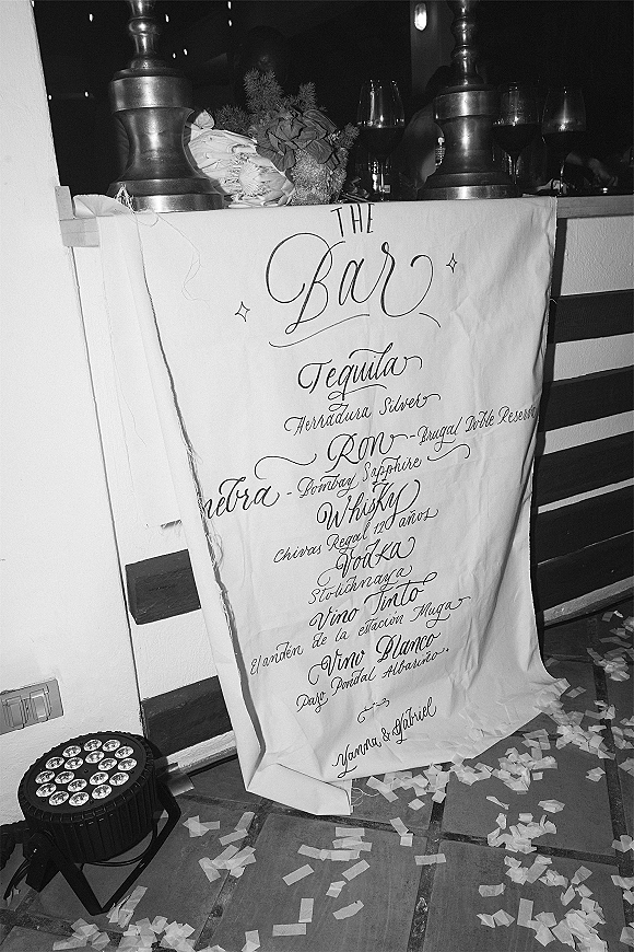 Wedding bar sign with calligraphy bar menu on hand-lettered fabric, tied with ribbon beside bottles, glasses, florals, and string lights