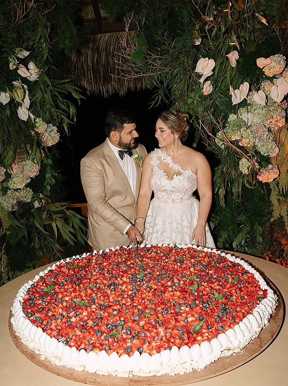 Wedding cake cutting by bride and groom beside an oversized sheet cake with mixed berries, under a floral arch at a nighttime dessert table