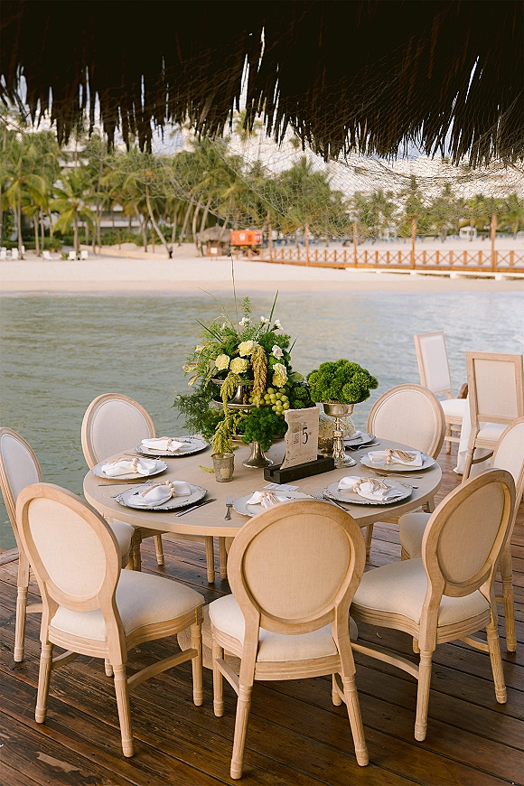 Reception tablescape on a round wedding table with green-and-white centerpiece, grapes and silver compotes, set by a dock with palm trees and water