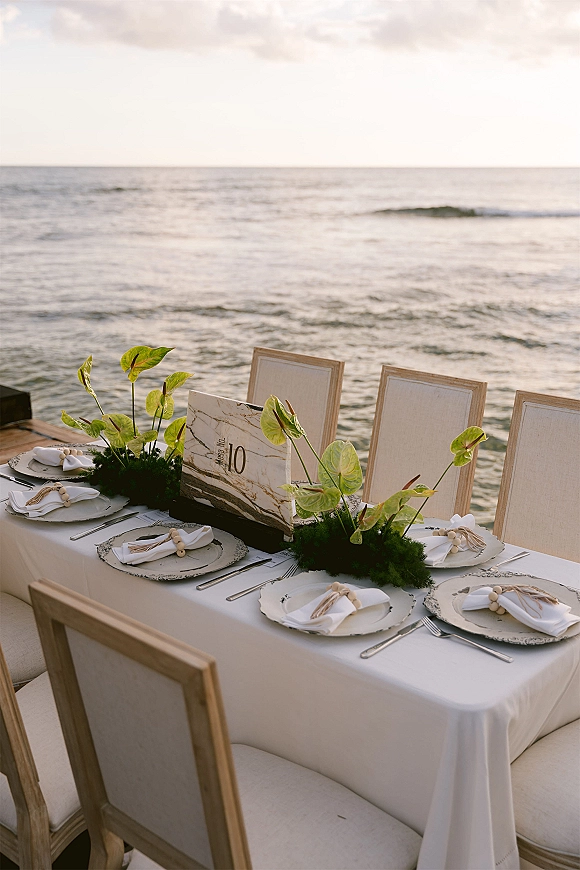 Reception tablescape with a beach wedding tablescape featuring white linens, ceramic plates, green anthurium centerpiece, and ocean horizon backdrop