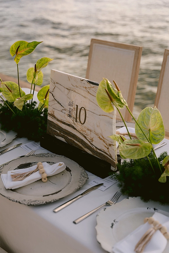 Reception tablescape wedding table setting with marble table number sign, moss runner, anthurium greenery, and waterfront backdrop