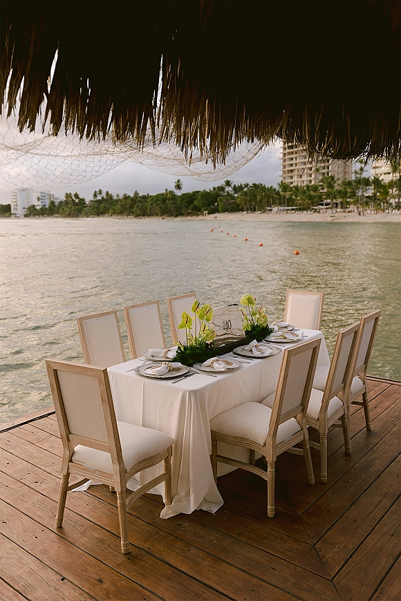 Reception tablescape with white linen, upholstered chairs, and a low anthurium centerpiece on a wood deck overlooking the ocean and palms