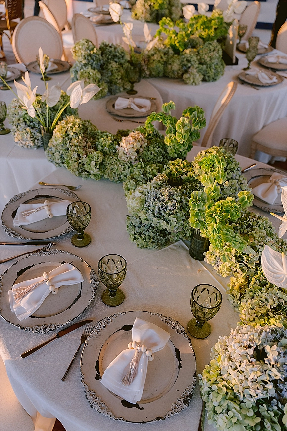 Reception tablescape with hydrangea garland centerpiece, green glass goblets, patterned plates, and white anthurium on white tablecloth indoors