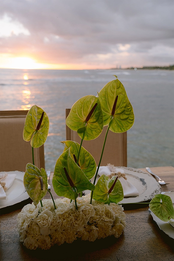 Reception tablescape with anthurium centerpiece and white flowers on a wooden table, place settings facing an ocean sunset under clouds