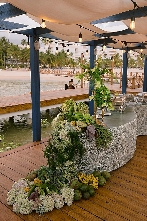 Wedding cocktail bar under string lights with fabric canopy, gold candelabra, greenery and hydrangeas on a beachfront resort deck by the water
