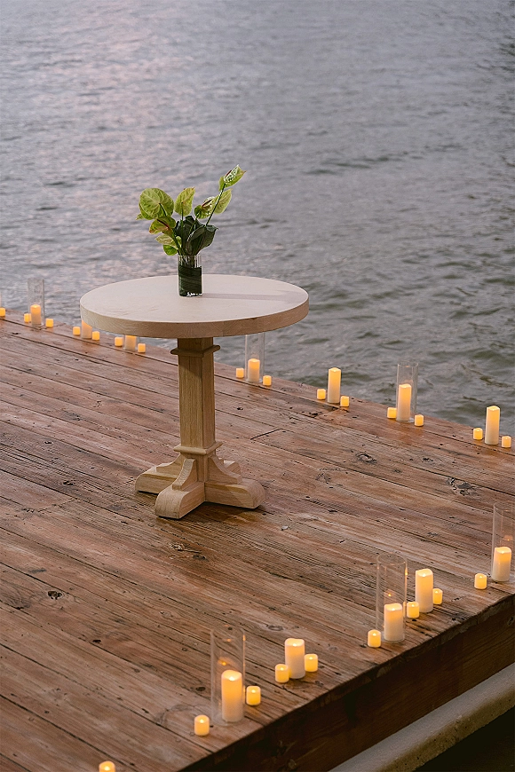 Ceremony setup for a dock wedding ceremony with round pedestal table, small floral vase, and pillar candles in hurricane vases on a lakeside dock