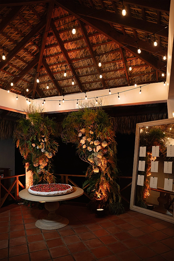Wedding dessert table with a wedding berry tart on a round pedestal, framed by hanging greenery, string lights, and a mirror seating chart display