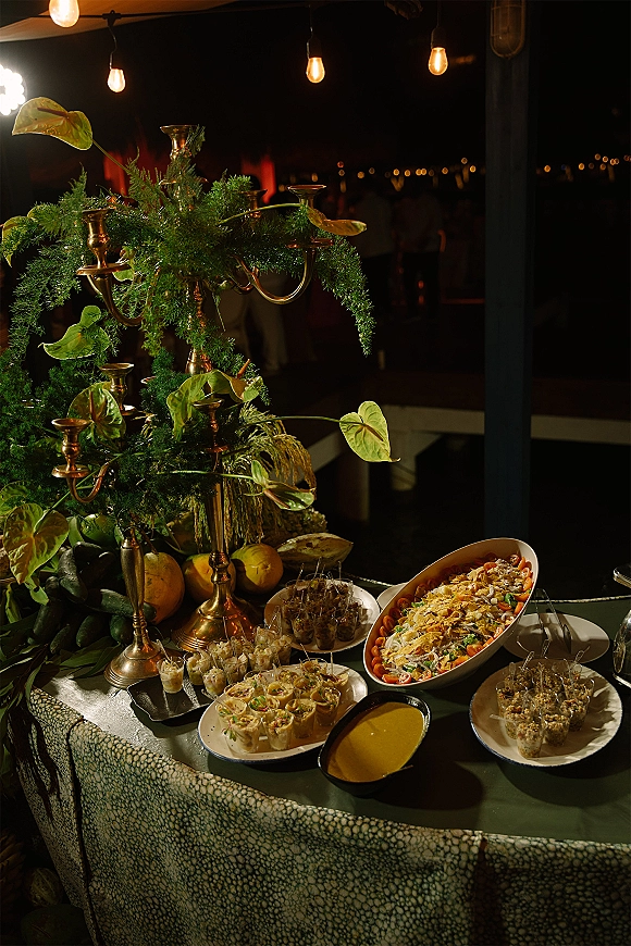 Wedding food display on a wedding buffet table with appetizer cups, fruit, salad and sauces, gold candelabra, greenery garland, string lights in a dark reception space