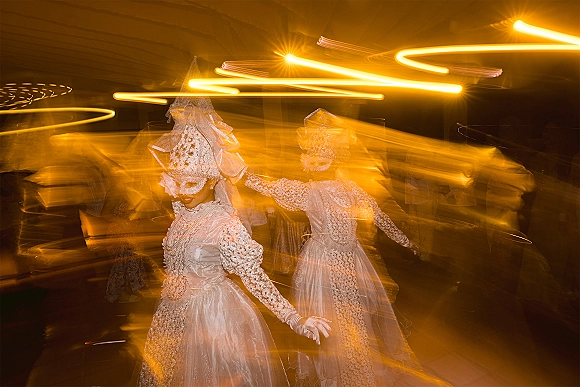 Wedding dance moment of brides twirling in embellished gowns with bridal veil headpiece, amid warm ceiling lights and guest silhouettes indoors