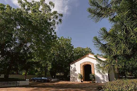 Wedding venue exterior with floral arrangements framing a brick arch doorway in a tree-lined courtyard, with a vintage convertible nearby