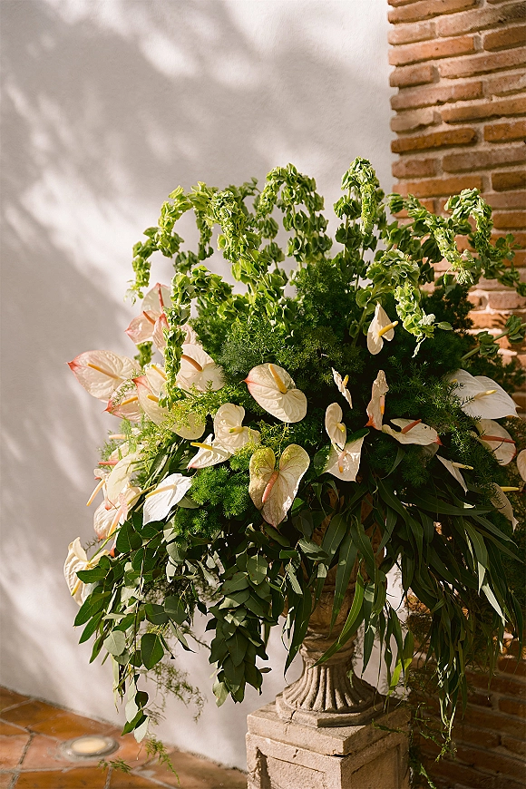 Wedding floral arrangement of white anthurium with trailing greenery in a pedestal urn beside a stucco wall and brick column