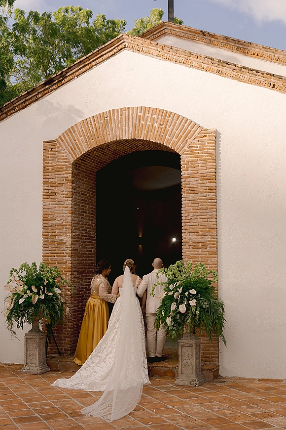 Wedding processional with bride walking into church from behind, long bridal veil trailing on terracotta tiles beneath a brick archway
