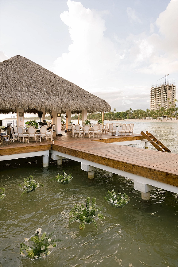Waterfront reception setup with white chairs and banquet tables under a thatched palapa on a dock, greenery centerpieces by the ocean