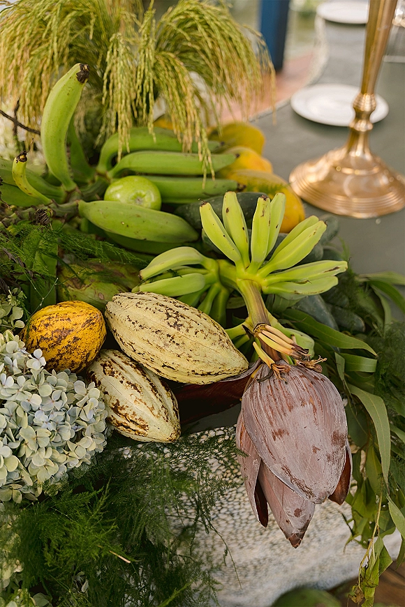 Fruit wedding centerpiece with tropical fruit, bananas and cacao pods nestled in greenery garland beside a brass candlestick on reception table