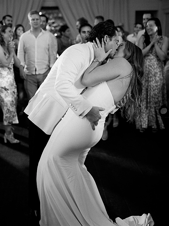 Wedding kiss during a first dance kiss as the groom dips the bride in a strapless dress on a reception dance floor with guests watching