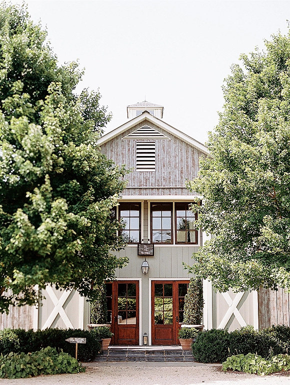 Barn wedding venue with rustic wedding venue exterior featuring wood double doors, lantern sconces, and potted topiary by a gravel path
