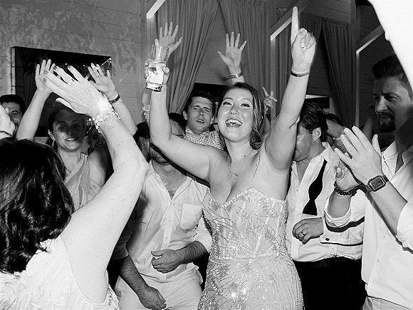 Wedding dance floor packed with guests cheering hands up as the bride in a strapless beaded dress dances holding a cocktail glass indoors