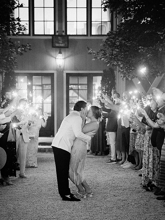 Wedding kiss portrait of bride and groom in a sparkler exit wedding tunnel as guests line a gravel courtyard with phones outside a windowed facade
