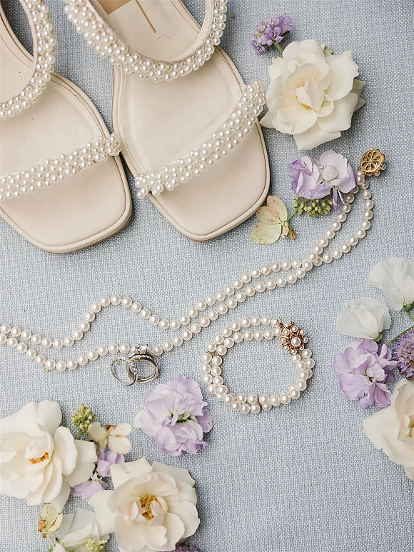 Wedding details flatlay with bridal accessories flatlay of pearl strap sandals, rings, pearl jewelry, flowers, and light blue fabric on textured backdrop