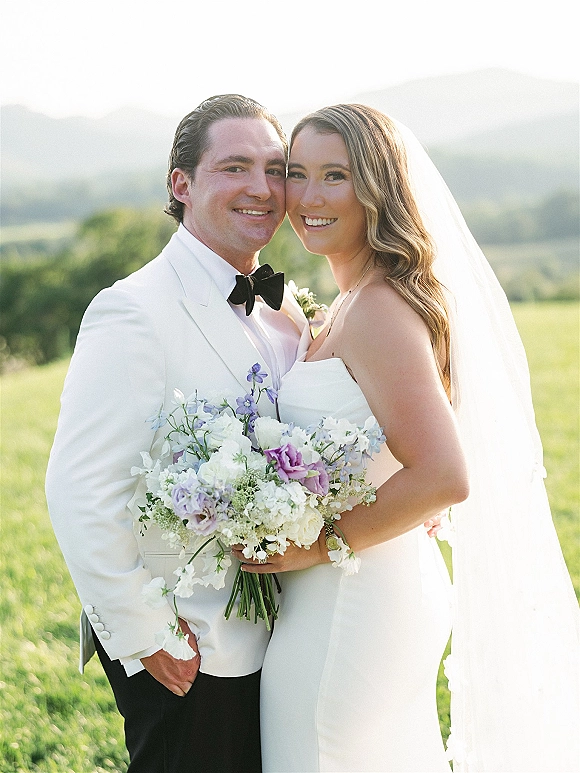 Couple portrait of bride and groom smiling, with lavender bouquet and long veil, set against green hills and mountains under sky