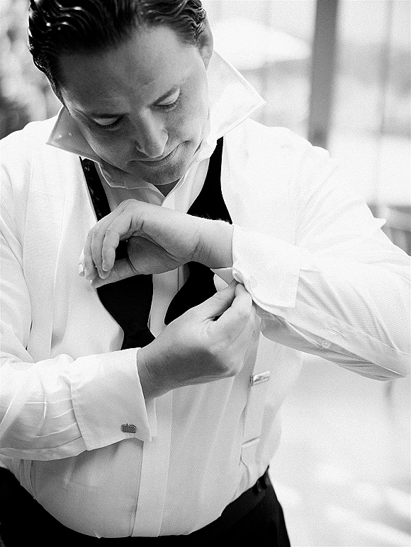 Groom getting ready, adjusting cufflinks on a white dress shirt by window light in an indoor room, with bow tie and waistcoat nearby