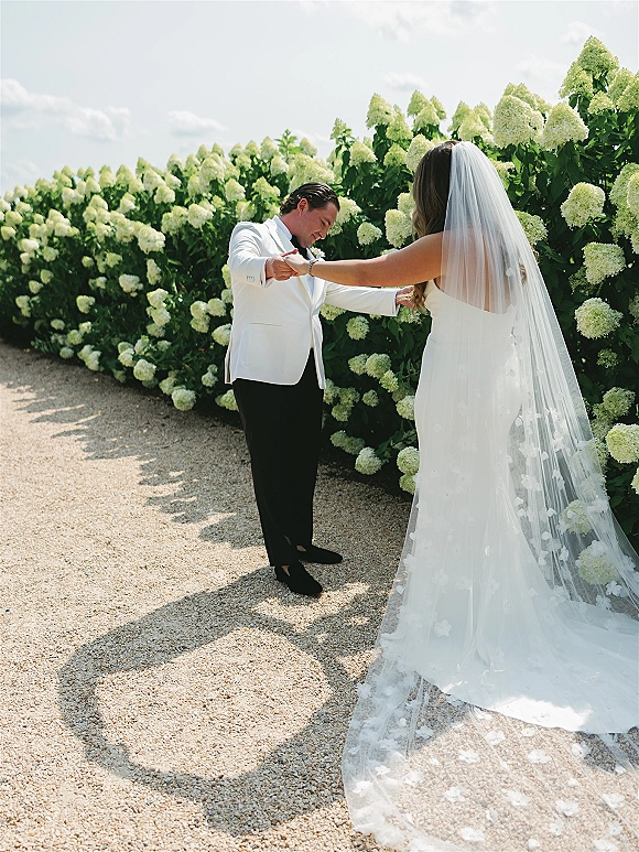 Couple portrait of bride and groom holding hands, her long veil and strapless dress flowing as he wears a white jacket by hydrangeas.