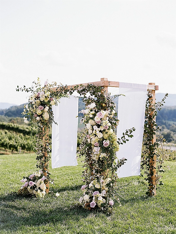 Wedding altar decor with floral ceremony arch of white drapery, roses, and hydrangeas on a vineyard lawn with distant hills